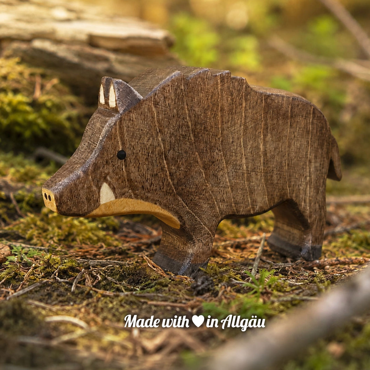 Handgefertigtes Holzspielzeug Wildschwein Keiler von Allgäuer Holztiere, auf einem moosbedeckten Waldboden vor einem Baumstamm stehend, umgeben von natürlichen Waldmaterialien, im Hintergrund unscharfe Waldlandschaft, aus bayerischem Ahornholz gefertigt, mit pflanzlichen Farben braun-schwarz. hellbraun und schwarz bemalt, im Allgäu produziert und für Kinder ab 3 Jahren geeignet.