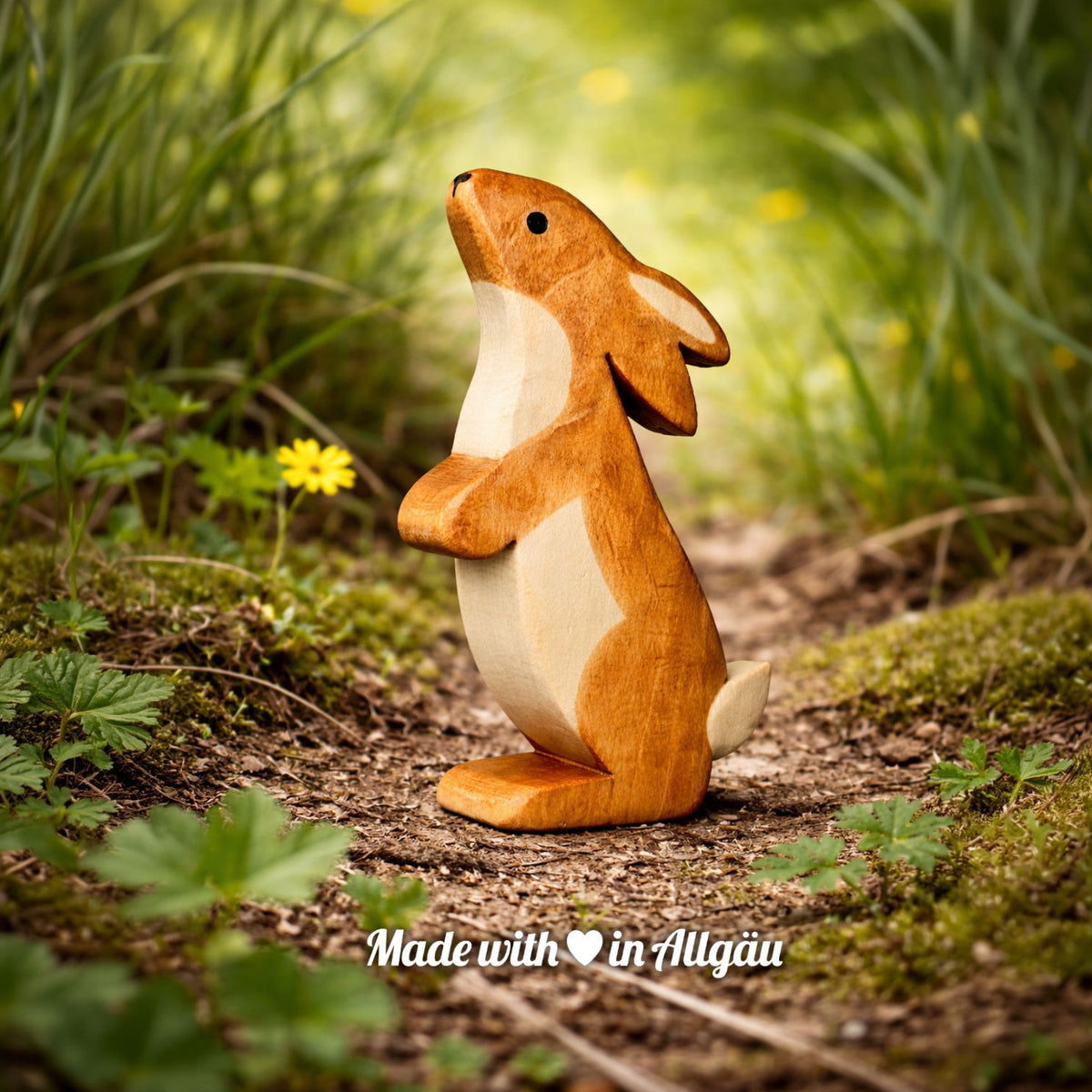 Handgefertigtes Holzspielzeug Feldhase  Rammler  Männchen von Allgäuer Holztiere, auf einem Waldweg zwischen Gras und kleinen Blumen stehend, im Hintergrund unscharfe grüne Vegetation, aus bayerischem Ahornholz gefertigt, mit pflanzlichen Farben braun und schwarz bemalt, im Allgäu produziert und für Kinder ab 3 Jahren geeignet.